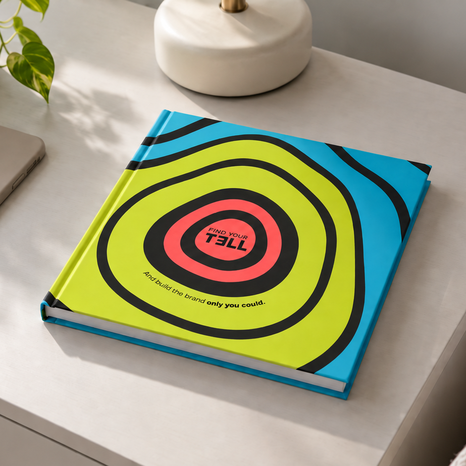 A hardcover Find Your Tell journal with a colorful concentric target design in lime green, yellow, pink, black, and blue, resting on a white bedside table next to a smartphone and a potted plant.