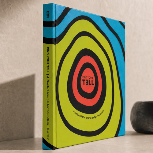 A hardcover Find Your Tell journal standing upright on a tabletop, featuring a bold concentric target design in bright yellow-green, black, blue, and coral red. The spine reads 'FIND YOUR TELL | A Guided Journal for Founders and the cover includes 'FIND YOUR TELL' with 'TELL' reversed as a play on words.